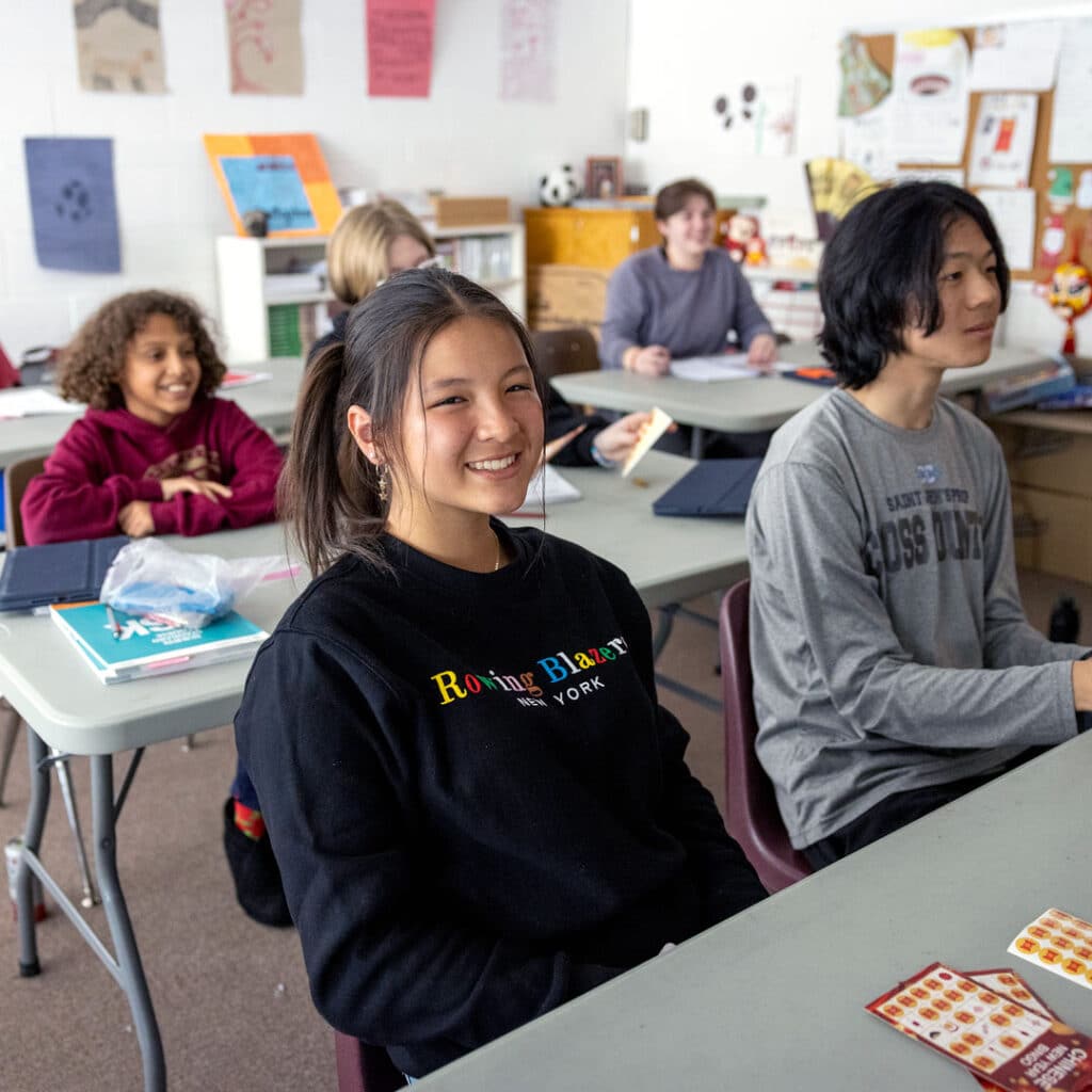 Prep Student In Sweatshirt Smiles At Camera While Classroom Of Math Students Learn In The Background